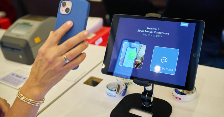 Image of a hand holding up a mobile phone, hovering over an iPad screen mounted on a stand offering self check-in at annual conference.